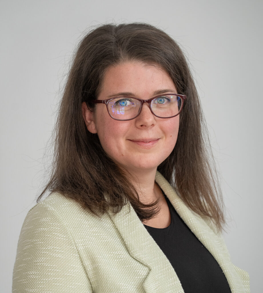 Fiatal, váll alatt érő barna hajú, szemüveges nő, világító kék szemekkel, kedves mosollyal. Drapp kosztüm kabátot, alatta fekete felsőt visel. félig a kép jobb oldala felé fordulva áll, de arcát a néző irányába fordítja.
