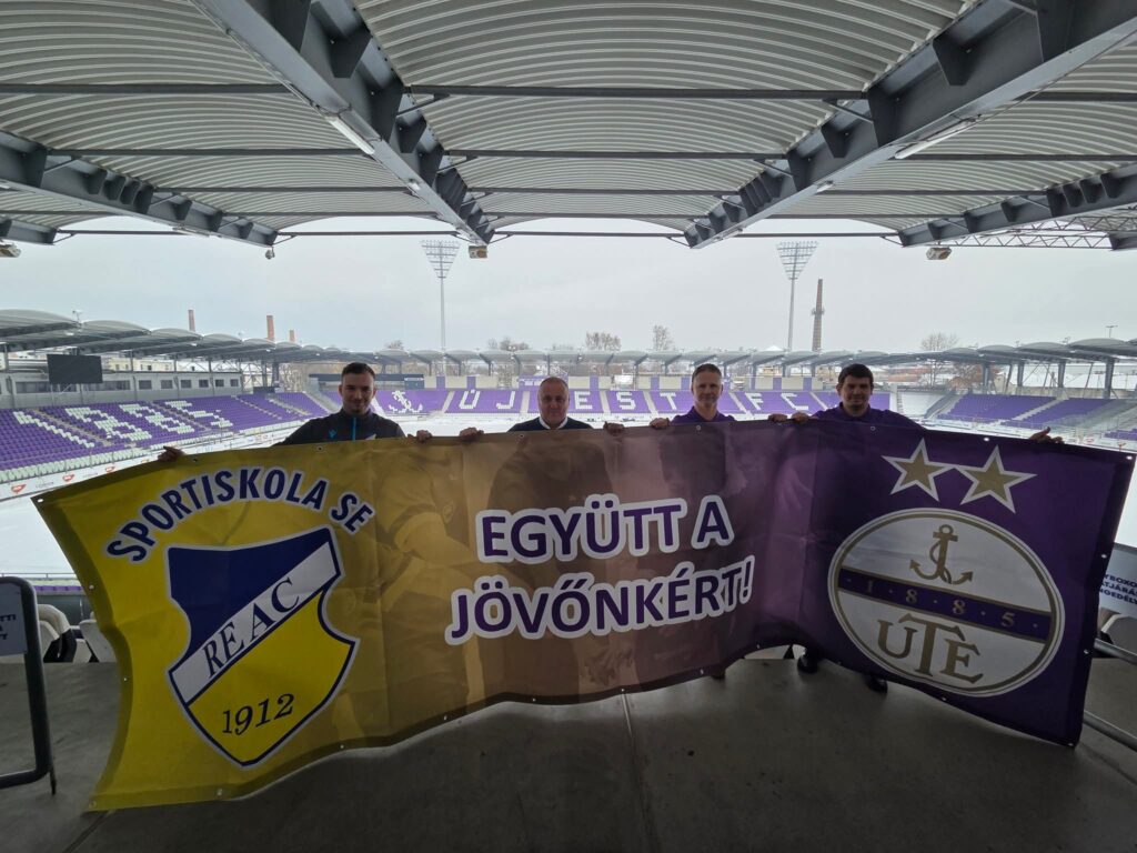 Négy férfi áll az Újpest FC stadionjának fedett lelátóján, és közösen tartanak egy nagy transzparenst. A banner bal oldalán a REAC Sportiskola SE sárga-kék logója látható „1912" évszámmal, jobb oldalán az UTE lila-fehér emblémája csillagokkal és „1885" évszámmal, középen pedig az „Együtt a jövőnkért!" felirat olvasható. A háttérben a havas pályával és lila széksorokkal teli stadion látható. A transzparens a két klub együttműködési megállapodását jelképezi.