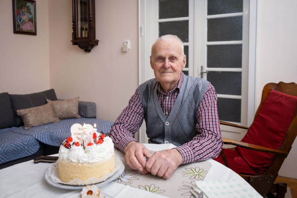 Gódor Antal, a 90 éves ünnepelt otthonának nappalijában egy asztalnál, előtte égő gyertyákkal és „Boldog születésnapot!” felirattal díszített fehér krémes születésnapi torta. Kockás inget és szürke mellényt visel, nyugodtan, méltósággal néz a kamerába. A háttérben kanapé, antik falióra és egy üvegajtó látható.
