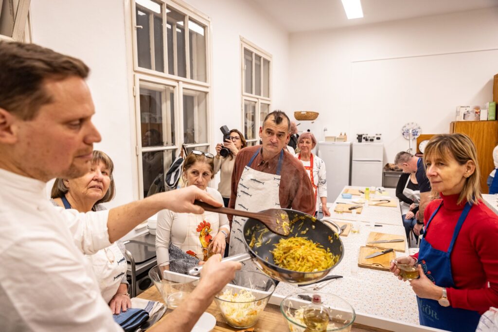 Egy világos, tágas konyhában a séf egy serpenyőt tart a kezében, benne pirított zöldségekkel, és éppen bemutatja a következő főzési lépést. A résztvevők kötényben állnak körülötte, figyelmesen követve a magyarázatot. A pultokon vágódeszkák és tálak sorakoznak, a háttérben nagy ablakok és konyhai berendezések láthatók. A jelenet a Ízutazás Pestújhelyen – Kulináris kalandok Kiszel Tamással program egyik aktív, tanulással teli pillanatát mutatja.