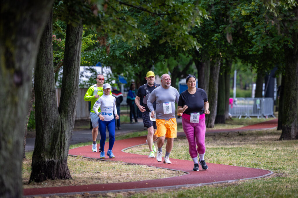 A fotón egy futóverseny résztvevői haladnak a Páskom park vörös burkolatú futópályáján, amely enyhén kanyarodva vezet végig a fás, zöld környezeten. Az előtérben egy mosolygó nő fut fekete pólóban és élénk rózsaszín csíkos leggingsben, mellette egy szürke felsőt és narancssárga rövidnadrágot viselő férfi halad, mindketten rajtszámmal a derekukon. Mögöttük további versenyzők követik őket különböző színű sportöltözetben, köztük egy fehér sapkás nő kék nadrágban és fehér felsőben, valamint egy neonsárga felsőt viselő férfi. A pályát kétoldalt magas fák szegélyezik, lombjuk árnyékot vet az útra, a háttérben pedig korlátok és a rendezvényhez kapcsolódó elemek láthatók. A résztvevők eltérő életkorúak, összpontosítva, ugyanakkor jókedvű hangulatban teljesítik a távot, a kép összességében a közösségi sportolás és az aktív életmód élményét közvetíti.