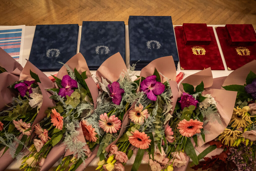 Egy terített asztalon több csokor friss virág és egymásra rendezett, díszes oklevélmappák láthatók. A virágcsokrok rózsaszín gerberákat, lila orchideákat és zöld leveleket tartalmaznak, halvány rózsaszín papírba csomagolva. Mögöttük két nagyobb halomban kék, mellettük kisebb számban bordó oklevélmappák sorakoznak, mindegyiken arany színű címerrel. A kompozíció ünnepi hangulatot áraszt, és egy közelgő kerületi elismerés vagy díszpolgári díjátadó előkészületeit idézi.