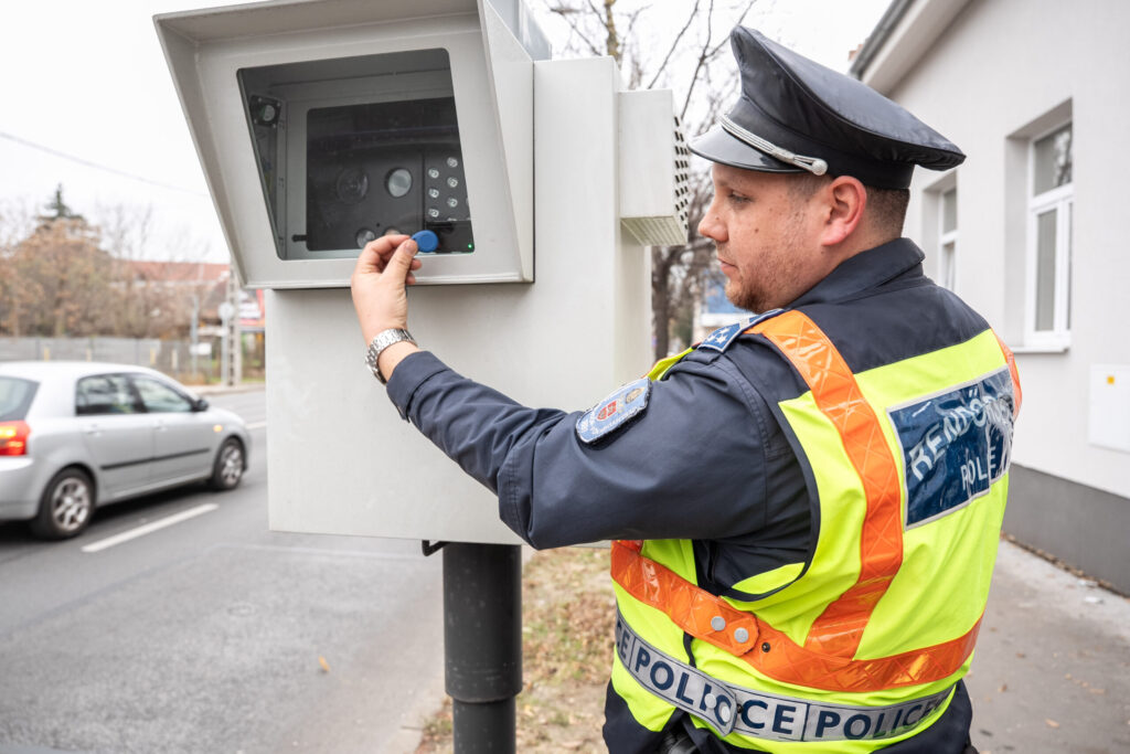 Egy rendőr egyenruhában egy fix traffiboxot állít be az út szélén. A mérőeszköz mögött elmosódva látszanak a nagy sebességgel haladó autók, érzékeltetve a forgalom gyors tempóját.