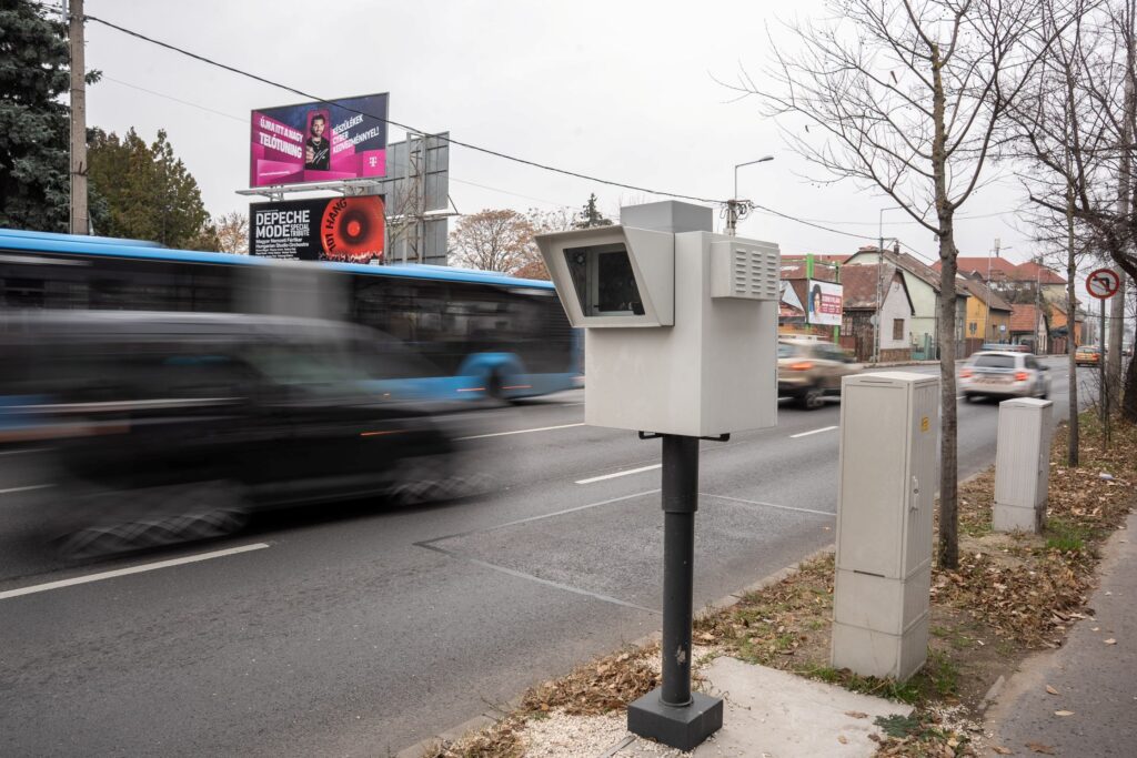 A fotón egy út mentén álló, oszlopra szerelt traffibox látható, amely egy forgalmas városi útszakaszt figyel. A kamera fekete tartóoszlopon áll, a háttérben több gyorsan elhaladó jármű – köztük egy kék busz és személyautók – mozgásuk miatt enyhén elmosódva jelennek meg. Az út túloldalán piros tetős lakóházak, kopasz fák és több utcai hirdetőtábla látható. A hirdetéseken különböző színes plakátok szerepelnek, köztük egy koncertplakát és egy magentaszínű szolgáltatói reklám. A jelenet téli vagy kora tavaszi hangulatú, a fények hidegek, a forgalom élénk, a traffibox pedig a közlekedés biztonságát szolgáló eszközként jelenik meg a kép középpontjában.