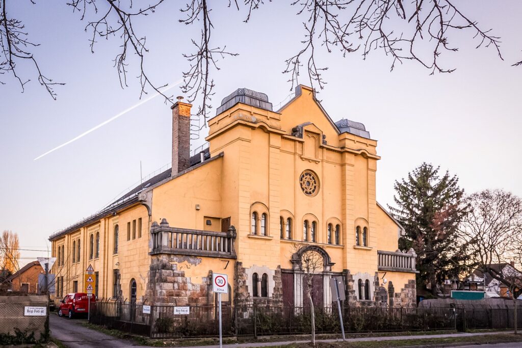 A fotón az impozáns, történelmi zsinagóga épülete látható Rákospalotán. A homlokzat világos, sárgás árnyalatú vakolattal készült, alsó része pedig kőburkolatot kapott. A főbejárat fölött díszes kőkeretezés és íves architektúra jelenik meg. A homlokzat felső részén egy nagy, kör alakú ablak látható, közepén Dávid‑csillag motívummal, amely a zsinagóga eredeti funkciójára utal. Az épület jobb oldalán egy magas, téglából épült kémény emelkedik. A tető peremén fémkorlát fut végig. A környezet csendes, városi utcarészlet: a kép előterében közlekedési táblák láthatók, köztük egy 30 km/h sebességkorlátozó tábla és egy utcanévtábla, amely a „Régi Főút” feliratot viseli. A jobb oldalon egy piros jármű parkol. A kép felső részét kopasz faágak keretezik, ami arra utal, hogy a fotó késő őszi vagy téli időszakban készült.