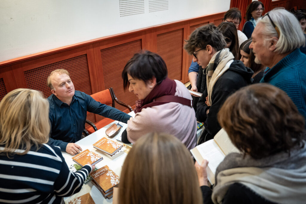 A fotón egy könyvbemutató és dedikálás pillanata látható a Csokonai Rendezvényházban. A kép középpontjában Lackfi János ül egy asztalnál, előtte több, egymásra rakott példány a frissen megjelent könyvéből. A borítók barna árnyalatúak, egységes megjelenésűek. Az asztal körül több érdeklődő áll, kezükben könyvekkel – némelyik nyitva, mások dedikálásra várva. A résztvevők beszélgetnek az íróval, vagy türelmesen várják a sorukat. A háttérben vörös falburkolat és narancsszínű ülőpárnákkal ellátott székek láthatók, a tér hangulata bensőséges és kulturális eseményhez illően nyugodt. A jelenet a Magyar Kultúra Napjához méltó, közösségi, irodalmi találkozást mutat: az olvasók és az író közvetlen kapcsolatát, a személyes dedikálás élményét.