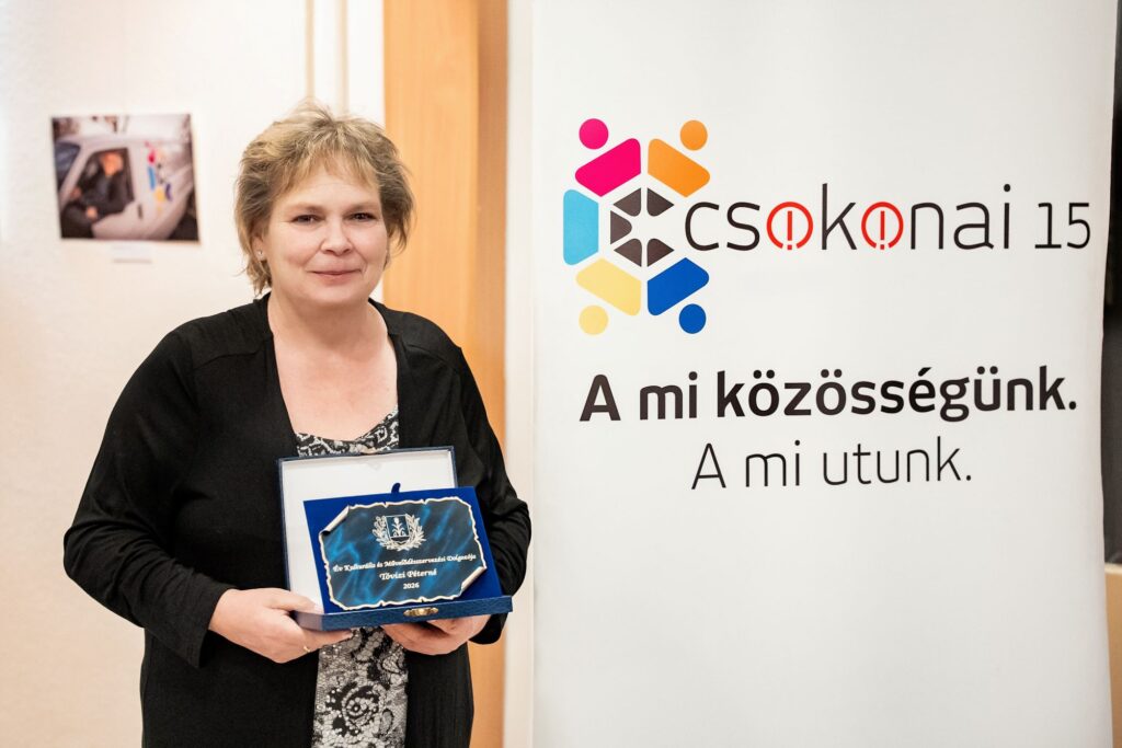 Tóvizi Péterné büszkén tartja kezében az „Év Kulturális és Művelődésszervezési Dolgozója 2026" feliratú díjplakettet kék bársonydobozban. Fekete kardigánban áll a Csokonai 15 reklámja előtt, amelyen „A mi közösségünk. A mi utunk." felirat olvasható.
