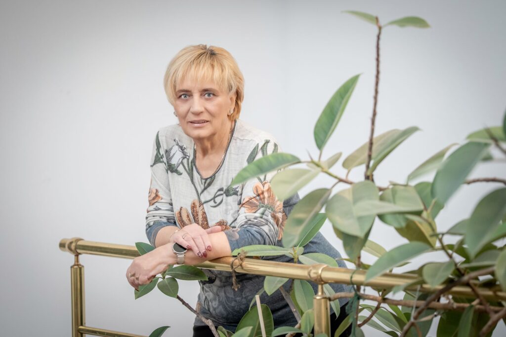 Tóth Vera képviselő félprofilból néz a kamerába, könyökét egy arany színű fémkorlátra támaszkodva. Rövid, szőke hajú, kék szemű, virágmintás szürke-kék felsőt visel, csuklóján okosórával. Előterében egy nagy levelű szobanövény látható.