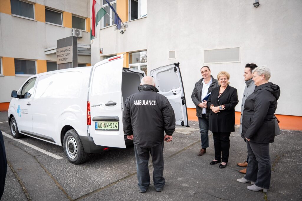 A fotón a XV. kerületi egészségügyi szolgálat számára átadott új elektromos autó bemutatása látható a Dr. Vass László Egészségügyi Intézmény előtt. A kép előterében öt ember áll egymás mellett: a kerület vezetőségének tagjai és az egészségügyi szolgálat munkatársai. A csoport mögött egy fehér, modern elektromos szolgálati jármű parkol, hátsó ajtói nyitva vannak. A jármű oldalán egészségügyi jelzés és intézményi felirat látható. A bal oldalon egy dolgozó áll, hátán jól olvasható az AMBULANCE felirat. Mellette a kerület vezetői sorakoznak: elegáns, hivatalos öltözetben, egyenes testtartással, az átadás ünnepélyes pillanatát tükrözve. A háttérben az egészségügyi intézmény bejárata látható, felette a felirat: „Dr. Vass László Egészségügyi Intézmény”. A homlokzaton több zászló is látható, köztük a magyar nemzeti zászló. A jelenet összhatása hivatalos, mégis közvetlen: a kerület vezetése egy új, környezetbarát jármű átadásával támogatja a helyi egészségügyi ellátást.