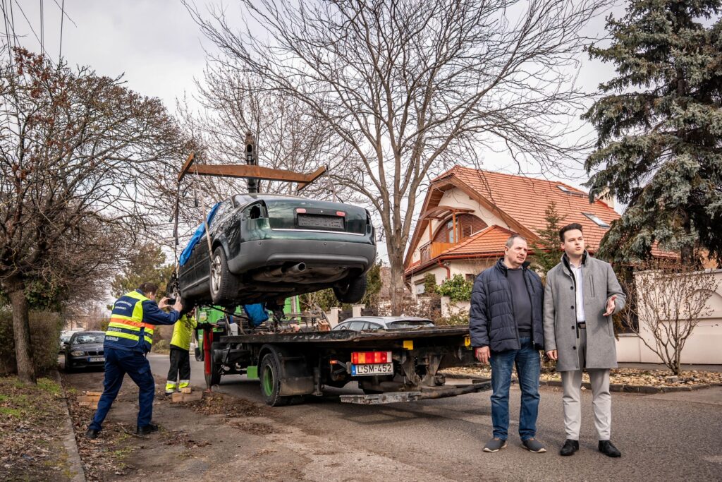 A fotón egy kertvárosias utcán zajló roncsautó‑elszállítási művelet látható. A kép középpontjában egy sötétzöld, erősen sérült és rendszám nélküli autó emelkedik a levegőbe, amelyet egy darus teherautó emel fel vastag hevederekkel rögzítve. A jármű láthatóan hosszabb ideje elhagyott állapotban volt. A teherautó mellett két láthatósági mellényt viselő munkatárs dolgozik: egyikük a daru működését felügyeli, másikuk a jármű stabilitását ellenőrzi. A háttérben családi házak, piros tetők, kopár fák és borús égbolt látható, ami hűvös időre utal. A kép jobb oldalán két, civil ruhás férfi figyeli a munkát: Makai Ferenc önkormányzati képviselő, Csernák János alpolgármester. Mindketten a kerületben zajló, felgyorsított roncsautó‑eltávolítási program helyszíni ellenőrzésén vesznek részt. Testtartásuk érdeklődő, a folyamatot közelről követik, jelezve, hogy a kerület vezetése kiemelten foglalkozik a közterületek rendezettségével és biztonságával. A jelenet a kerület azon törekvését mutatja be, hogy a közterületeken hagyott, veszélyes vagy elhanyagolt járművek mielőbb eltűnjenek, tisztább és biztonságosabb környezetet teremtve a lakóknak.