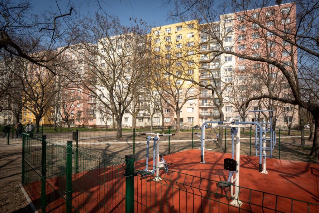 A fotón egy felújított, szabadtéri kondipark látható, amelyet piros gumiburkolat fed. A területet zöld fémkerítés veszi körül, belül pedig több, különböző izomcsoportokat megmozgató kültéri fitneszgép áll egymás mellett. A gépek fémből készültek, stabil, strapabíró kialakítással. A kondiparkot lombtalan fák övezik, ami arra utal, hogy a kép késő őszi vagy téli időszakban készült. A háttérben több többszintes, pasztellszínű társasház látható — sárga, rózsaszín és bézs árnyalatokkal —, amelyek között a napfény erősen megvilágítja a jelenetet. A park rendezett, tiszta, jól karbantartott, és azt sugallja, hogy a környék lakói számára egy modern, közösségi sportolási lehetőség vált elérhetővé.