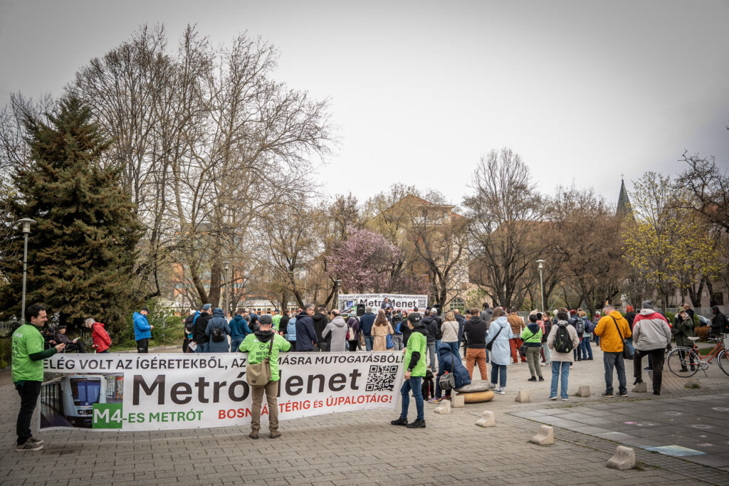 Civilek gyülekeznek egy városi téren a hiányzó metrószakasz megépítését támogató rendezvény előtt. A résztvevők egy nagyméretű transzparens körül gyűlnek össze, hogy közösen hívják fel a figyelmet a régóta várt közlekedési fejlesztés fontosságára.