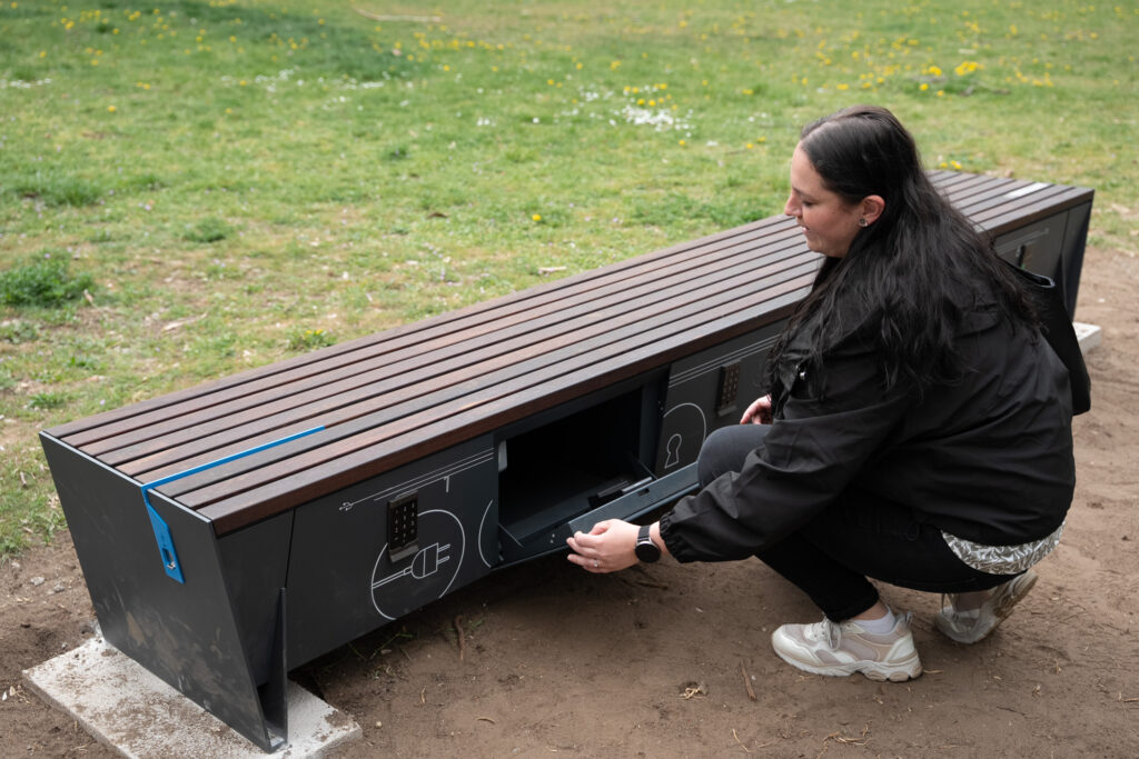 Csomagmegőrzés a parkban. A képen egy modern, fából készült okospad látható, amelynek oldalán zárható rekesz nyílik; egy személy éppen kinyitja a tárolófülkét a zöld parkterületen.