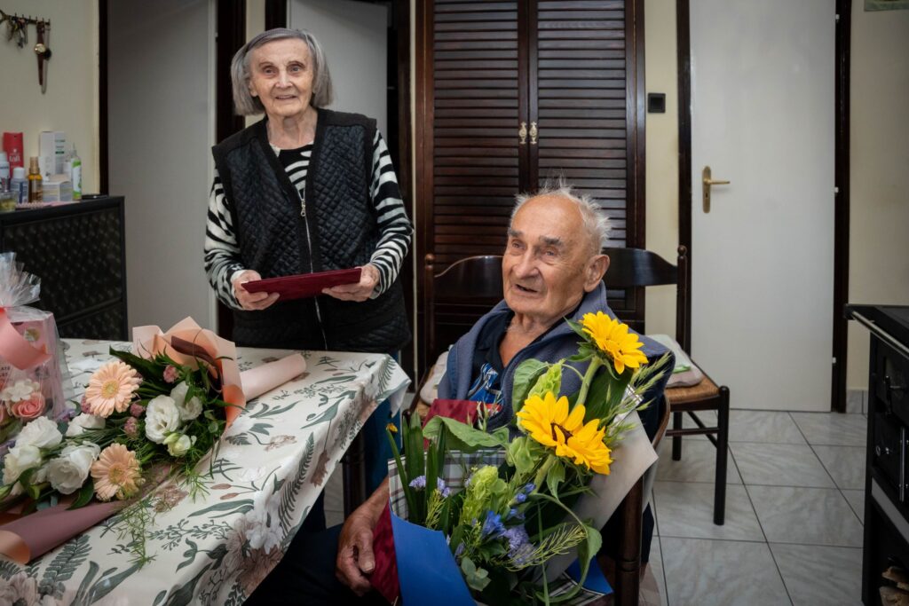 A rákospalotai Seitler–Monori házaspár együtt ünnepli a 90. és a 95. születésnapját. A képen az idős pár virágok és ajándékok között ül és áll, otthoni környezetben.