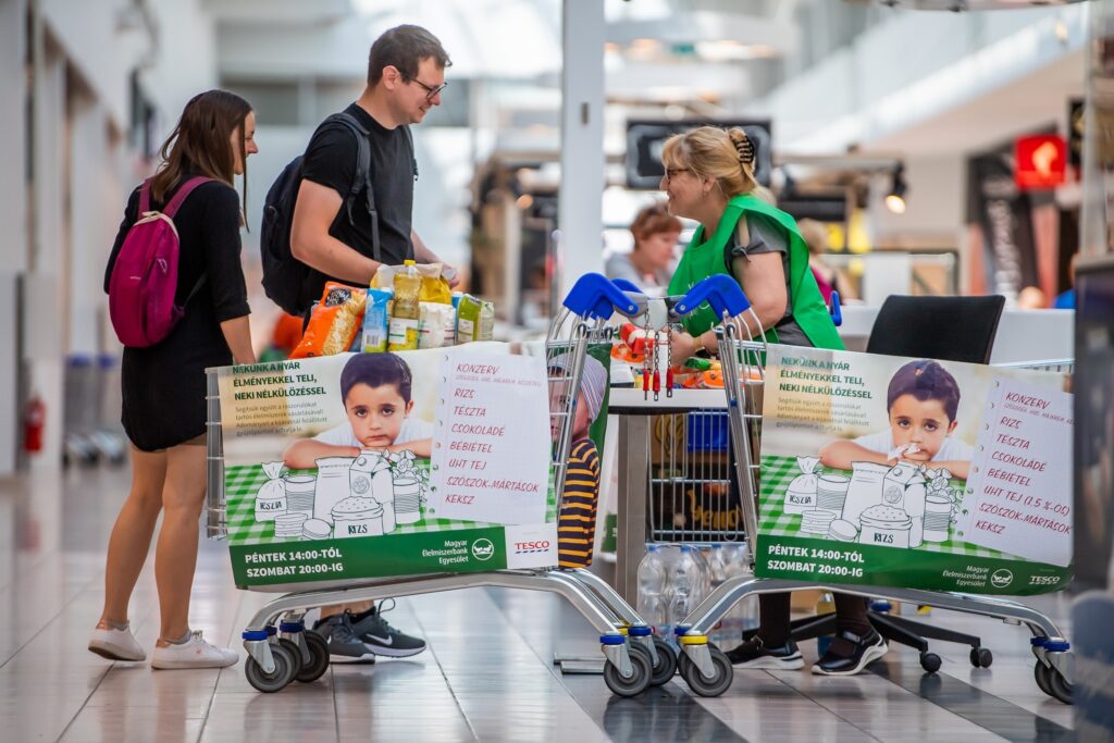 Önkéntesek az ország legnagyobb élelmiszer‑gyűjtő akcióján. A képen adományokkal teli bevásárlókocsik és a gyűjtést segítő önkéntesek láthatók egy áruház bejáratánál.