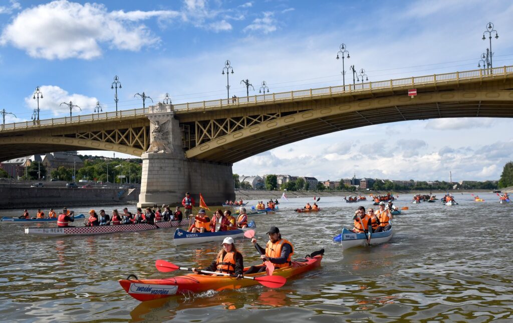 Budapest Átevezés. A képen narancssárga mentőmellényt viselő résztvevők kajakokkal és kenukkal eveznek a folyón, a díszes, ívelt Margit híd alatt, városi háttérrel.