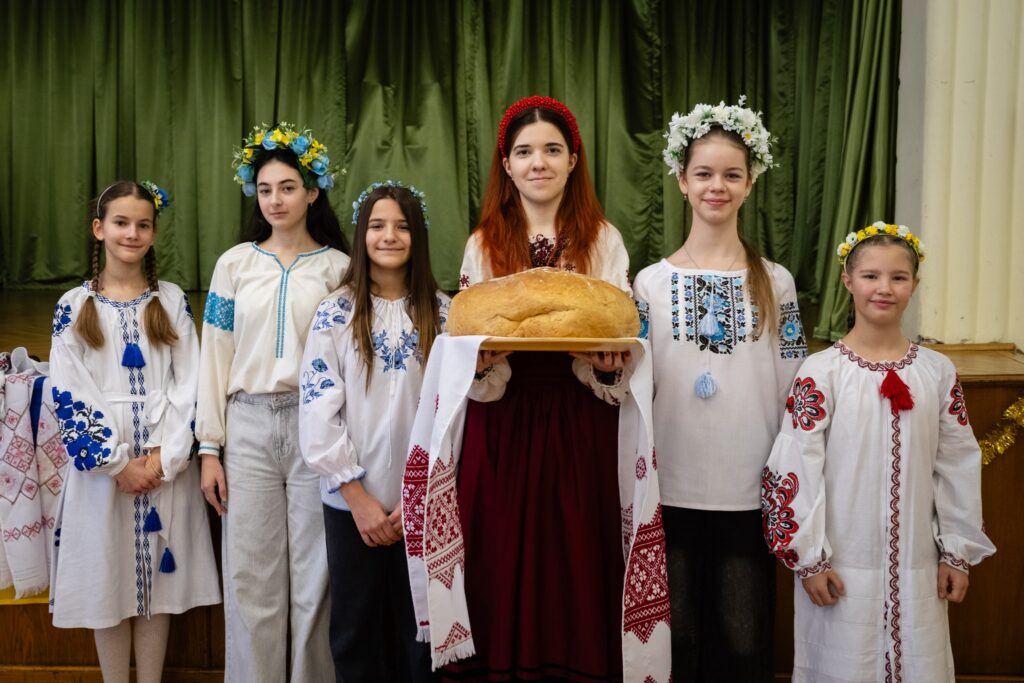 Hat lány áll egy zöld függönyös színpad előtt, mindannyian hagyományos ukrán hímzett blúzban – vyshyvanká-ban. Fejükön virágkoszorút viselnek. A középső, vörös hajú lány piros fejdíszszel és bordó szoknyával hímzett kötéssel díszített ruhában egy kerek fehér kenyeret tart tálcán, hímzett kenőre terítve – ez a hagyományos ukrán kenyér-só fogadtatás jelképe.