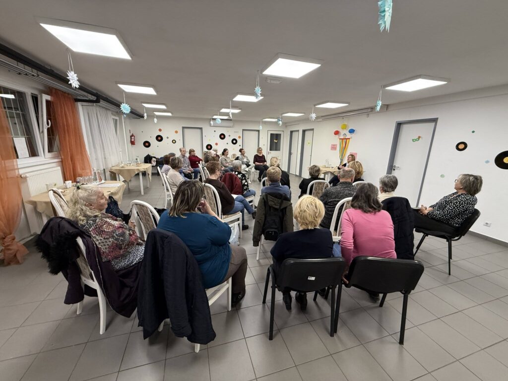 Alzheimer Café Újpalota. A képen egy világos, közösségi térben ülő csoport látható, ahol az érdeklődők előadást vagy beszélgetést hallgatnak, a terem falán színes dekorációkkal.