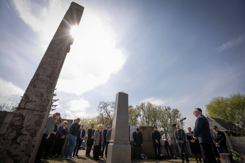A rákospalotai és pestújhelyi zsidó temetőben megemlékezést tartottak a holokauszt helyi áldozatairól. A képen a résztvevők egy kőemlékmű előtt állnak, miközben egy felszólaló mikrofon mögött beszédet mond a napsütéses, csendes környezetben.