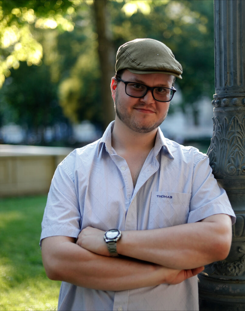 Fiatal, szemüveges férfi a szabadban, nyitott nyakú szürke ingben. Keki színű lapos, úgynevezett barett sapkát visel. Rövid, borosta jellegű szakálla és bajusza van, mosolyog.