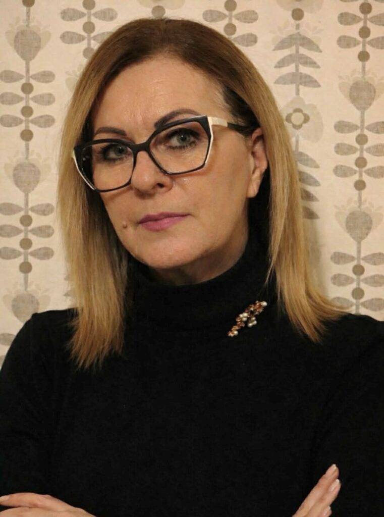 Szőke, vállig érő hajú, szabályos arcú szemüveges nő poirtréja. Fejét picit a kép bal oldala felé fordítja, de szembe néz. Sötét garbót visel.