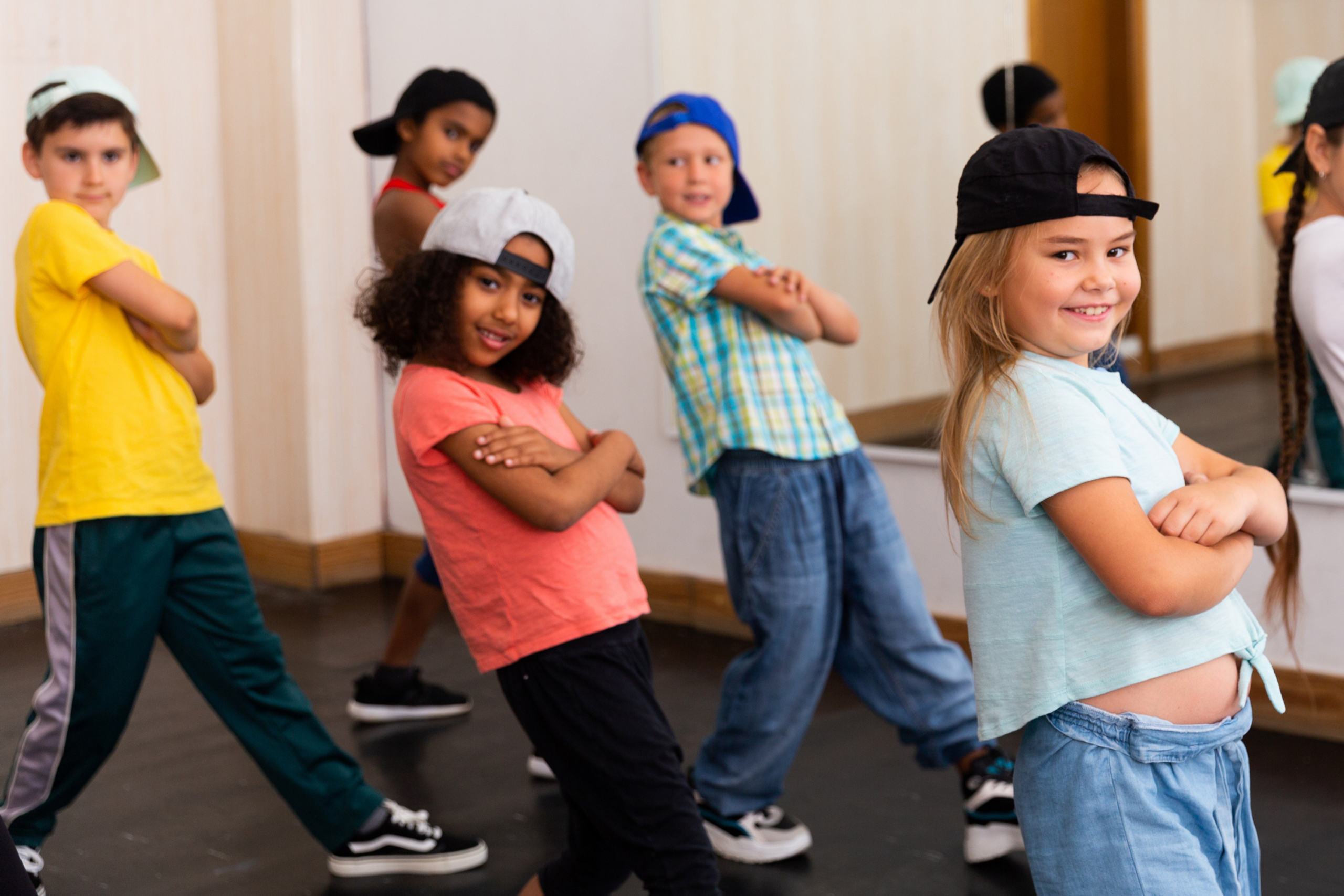 Hat gyermek baseballsapkában hiphopot táncol és mosolyog a kamerába. Mögöttük a falon egy nagy tükör látható.