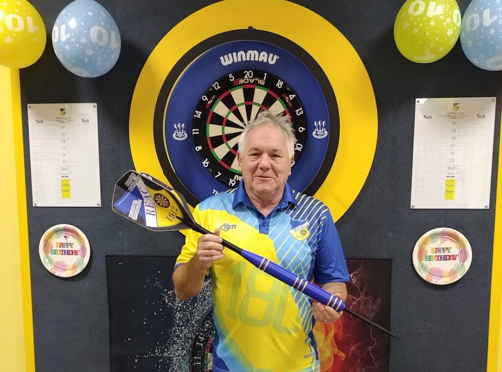 Handel Károly, a REAC Sportiskola darts szakosztályának egyik alapítója és technikai vezetője egy Winmau dartstábla előtt áll, sárga-kék REAC csapatmezben. Kezében a klub logójával és „REAC 1912" felirattal díszített, egyedi darts nyilakat tart. A tábla körül születésnapi lufikkal és „Happy Birthday” feliratú tányérokkal díszített a fal – nagy jubileumi vagy születésnapi alkalom keretében készült a fotó.