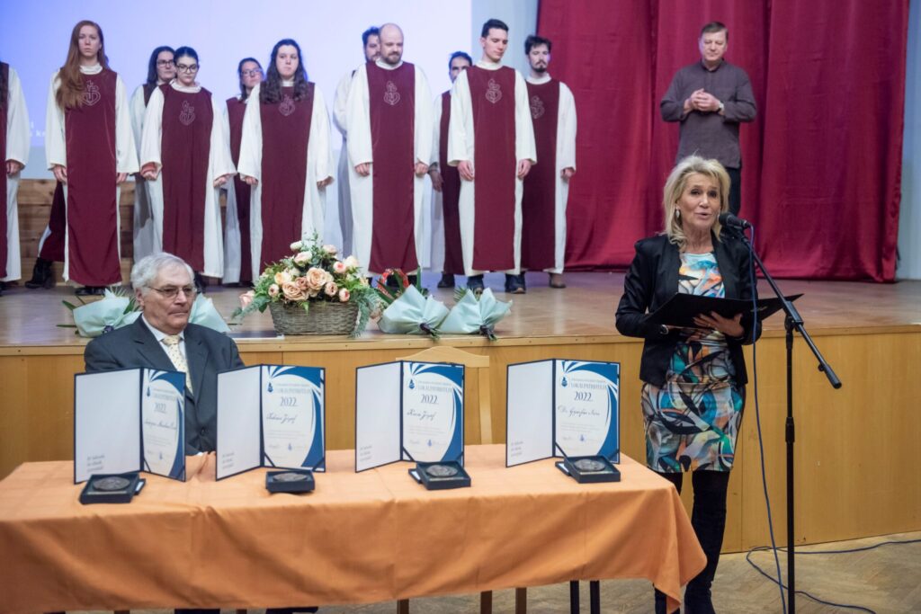 Lokálpatrióta díj átadása. Az asztalnál a Lokálpatrióták a XV. kerületért Egyesület elnöke, Németh Péter ül, a mikrofon előtt pedig Gressó Ildikó, a Polgármesteri Kabinet vezetője beszél. Az asztalon oklevelek és díjak láthatók, mögöttük kórus tagjai állnak sorban.