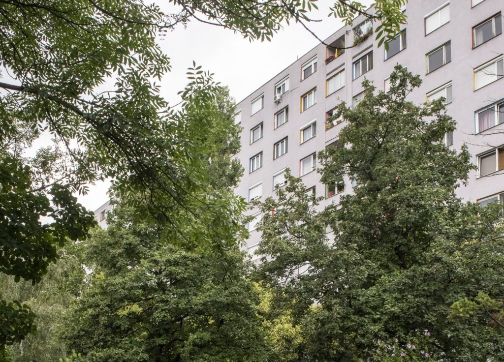 Egy többemeletes, világosszürke homlokzatú újpalotai lakóház látható, amelyet sűrű, zöld lombú fák takarnak részben az előtérben. Az épület hosszú, egyenes vonalú ablaksoraival tipikus lakótelepi társasház benyomását kelti. Néhány ablaknál függöny vagy növény látható, egyes erkélyeken virágok is megjelennek. A fák élénk zöldje és a ház világos színei nyári vagy késő tavaszi hangulatot sugallnak. A jelenet harmonikusan mutatja be, hogyan simulnak össze Újpalota lakóépületei a környező városi zöldfelületekkel.