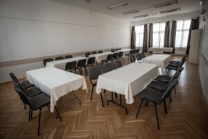 Hosszúkás, parkettás terem, körben terített asztalpkkal berendezve. A jobb oldali falon ajtó, a szemköztin ablakok, félrehúzott szürke sötétítő függönyökkel.