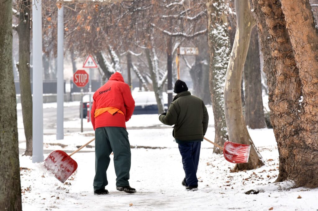 A fotón egy havas járdán halad végig két ember, akik piros hókotró lapátot visznek magukkal. Mindketten vastag, téli ruházatot viselnek. A bal oldalon lévő személy élénk piros kabátot hord, hátán jól látható a Steward felirat, hozzá zöld nadrágot visel. A mellette haladó társa sötétzöld kabátban és sötét nadrágban lépked. Mindketten előre néznek, határozott, céltudatos mozdulattal haladnak a havas úton. A járda két oldalán kopasz téli fák sorakoznak, ágaikon vékony hóréteggel. A háttérben egy STOP tábla és egy gyalogátkelőhelyet jelző tábla látható, ami városi környezetre utal. A levegő tiszta, hideg, a jelenet csendes téli reggelt idéz, ahol a közterület rendben tartásán dolgozók épp a hó eltakarítására készülnek.