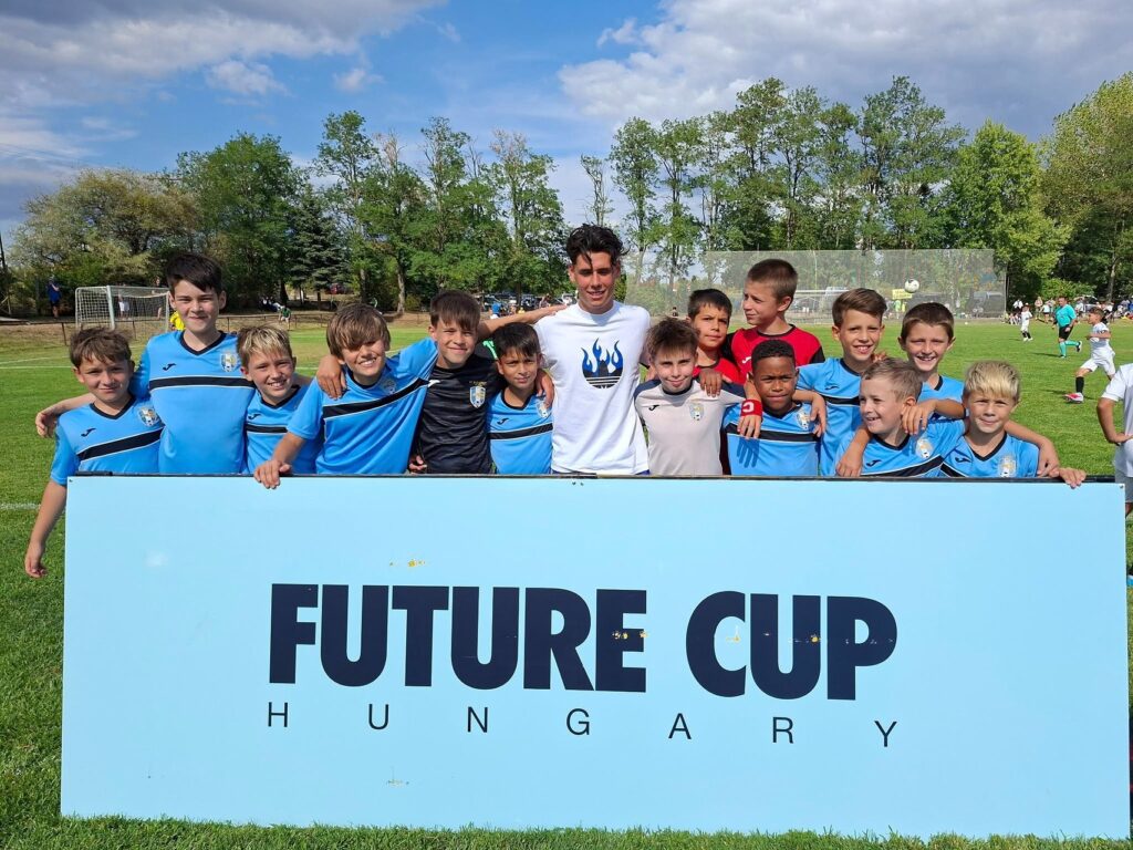 Egy futballpályán fiatal gyerekcsapat sorakozik fel egy „Future Cup Hungary” feliratú tábla mögött. A csoport közepén Skribek Alen Martin NB I-es labdarúgó áll fehér mezben, mosolyogva. A körülötte álló gyerekek többsége kék-fekete Joma csapatmezben van. A háttérben zöld fák, focikapu és további játékosok láthatók napos időben.