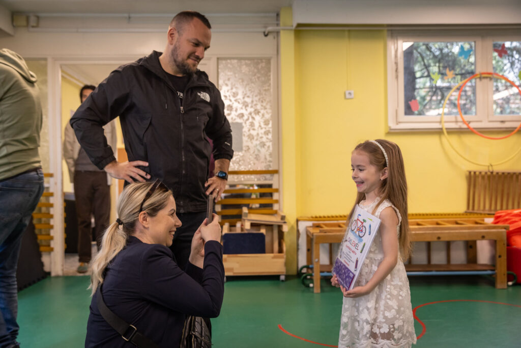 „Kerékpározni és rajzolni is jó” díjátadó. A képen egy kisgyermek áll oklevéllel a kezében egy színes, óvodai teremben, miközben egy felnőtt fényképet készít róla, mások pedig figyelik a pillanatot.
