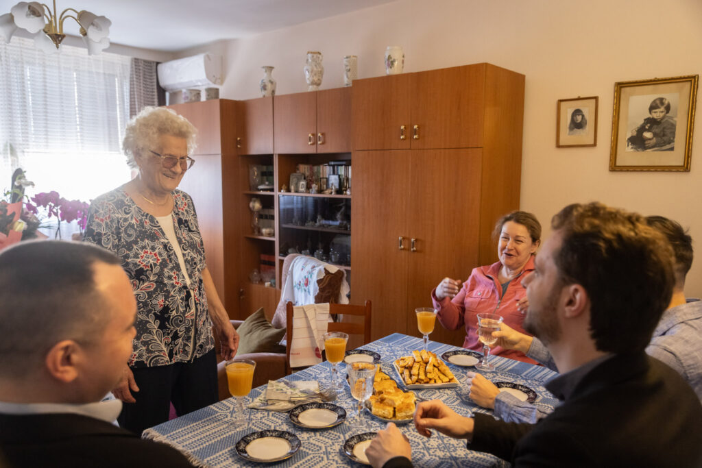 A fotón egy meghitt, otthoni környezetben zajló ünneplés látható. A jelenet egy világos, barátságos étkezőben játszódik, ahol a középen álló asztalt kék mintás terítő fedi. Az asztalon narancslé, tányérok, poharak és több tálca házi sütemény sorakozik, mintha vendégvárásra készültek volna. Az asztal körül öt ember foglal helyet vagy áll. A kép középpontjában egy idős hölgy látható, aki mosolyogva áll az asztal mellett – ő Juhász Jánosné Tóth Mária, aki a 90. születésnapját ünnepli. Körülötte a vendégek ülnek és beszélgetnek, oldott, barátságos hangulatban. A hölgyet személyesen köszönti: Gyurkó Dániel polgármester, Pósa András önkormányzati képviselő. Mindketten tiszteletteljes, ünnepi jelenléttel vesznek részt a köszöntésben, figyelmesen fordulnak az ünnepelt felé. A háttérben fa bútorok, vitrinek, családi fényképek és egy ablak látható, amelyen át természetes fény árad be, tovább erősítve a meghitt, családias hangulatot. A jelenet a kerület hagyományos idősköszöntő programjának része, amelyben a helyi vezetők személyesen látogatják meg a szépkorú lakosokat, hogy méltó módon ünnepeljék életük fontos mérföldköveit.