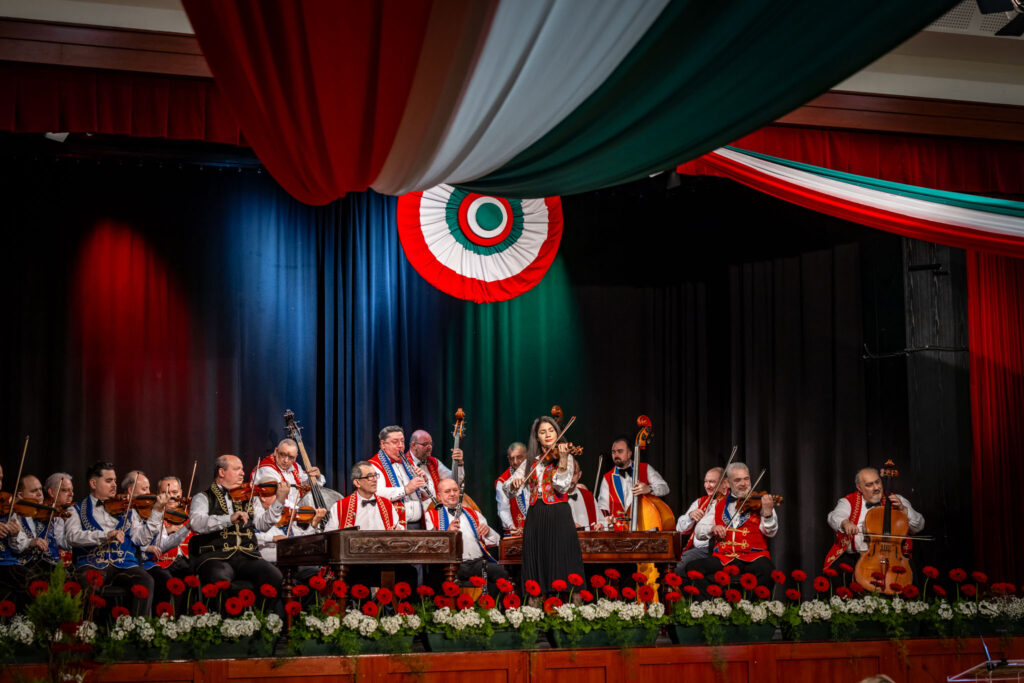 A március 15-i ünnepség kulturális műsorán a 100 Tagú Cigányzenekar teljes együttese látható a színpadon. A zenészek hagyományos, hímzett mellényekből és fehér ingekből álló fellépőruhát viselnek. A színpadot piros‑fehér‑zöld drapériák és virágdíszek keretezik, fölötte nagy, kör alakú nemzeti színű embléma függ. A zenekar tagjai vonós hangszereken – hegedűkön, brácsákon, csellókon, nagybőgőkön – játszanak, elöl két cimbalom is látható. A zenészek koncentráltan, összehangoltan adják elő műsorukat, a jelenet pedig ünnepélyes, emelkedett hangulatot áraszt, méltó keretet adva a nemzeti ünnepnek.