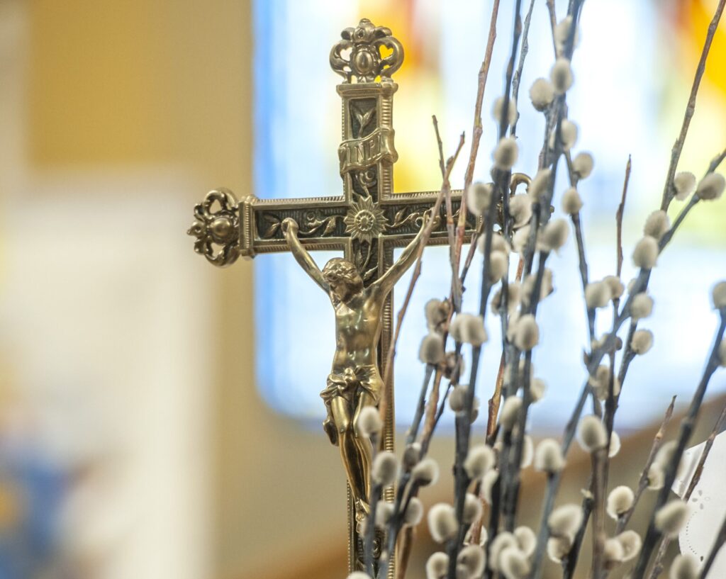 A fotó középpontjában egy díszes fém feszület látható, rajta a keresztre feszített Jézus alakjával. A kereszt előtt és mellett barkaágak emelkednek a kép előterében, puha, ezüstös-fehér barkarügyekkel. A háttér életlen, meleg színű templomi vagy kápolnai belső teret sejtet, ahol a beszűrődő természetes fény nyugodt, ünnepi hangulatot teremt. A barka a tavasz közeledését és a megújulást jelképezi, míg a feszület a keresztény húsvét központi üzenetére, Jézus szenvedésére, halálára és feltámadására utal. A kép a húsvétot mint a remény, az újjászületés és az élet győzelmének ünnepét idézi meg.