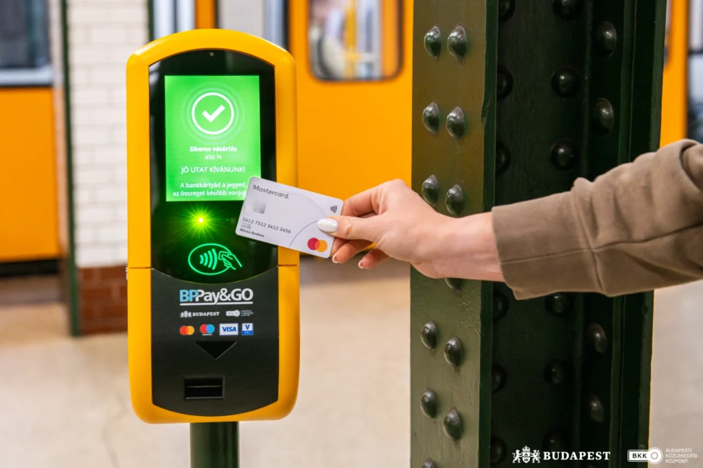 Közeli felvétel egy sárga színű, érintésmentes bankkártyás jegyvásárló és érvényesítő készülékről egy metróállomáson. A kép jobb oldalán egy kéz bankkártyát tart a készülék leolvasója elé. A kijelző zöld háttérrel világít, rajta pipa jelzi a sikeres tranzakciót, valamint a „Sikeres vásárlás” üzenet olvasható. A készülék alsó részén az érintésmentes fizetés szimbóluma és a BP Pay&GO felirat látható. A háttérben elmosódva egy sárga metrókocsi és az állomás szerkezeti elemei jelennek meg. A fotó a budapesti közösségi közlekedés digitális, bankkártyás jegyvásárlási lehetőségét szemlélteti.