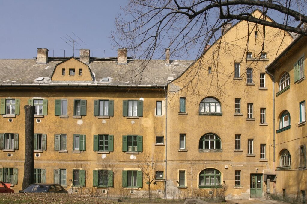 Egy többemeletes, régi építésű MÁV‑telepi lakóház látható. A homlokzat sárgás‑barnás vakolatú, több helyen kopott és foltos, ami az épület korát és időjárás okozta elhasználódását mutatja. A ház számos ablakán zöld spaletták vannak, némelyik nyitva, mások csukva. A tető szürke, több kéménnyel és néhány tetőablakkal. Az előtérben két lombtalan fa áll, mellettük kis füves terület és két parkoló autó. A jelenet késő téli vagy kora tavaszi hangulatot áraszt, a fák csupasz ágai és a tompa fény miatt.
