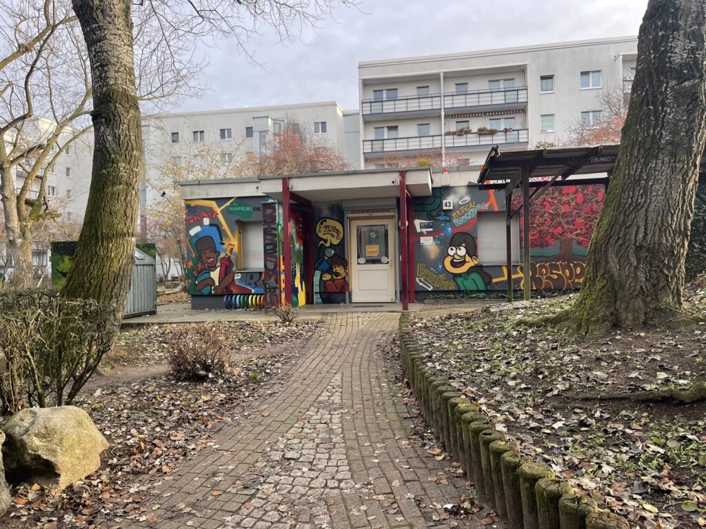 Egy alacsony, egyemeletes épület látható parkos környezetben, őszi lombtalan fák között. Az épület teljes homlokzatát színes, rajzfilmszerű graffiti falfestmények borítják, melyeken figurák, feliratok és egy fa motívuma látható. A bejárathoz téglajárda vezet, a kert tele van lehullott falevelekkel. A háttérben egy panelház emelkedik. Az épület érdeklődés ifjúsági ház.