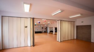 Egy terem, amelynek szemközti oldala félig nyitott harmonika ajtó szerű válaszfal. a fény baloldalról érkezik, de az ablak nem látszik. jobra nagy, 2 szárnyas ajtó.