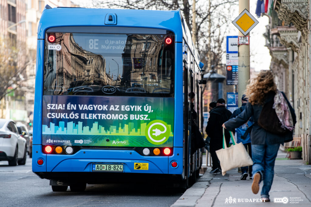 Egy kék, csuklós elektromos autóbusz áll a megállóban, ahol utasok szállnak fel. A jármű hátulján jól látható a 8E járatszám és az Újpalota felé tartó úticél, valamint a város új elektromos buszait bemutató grafika.