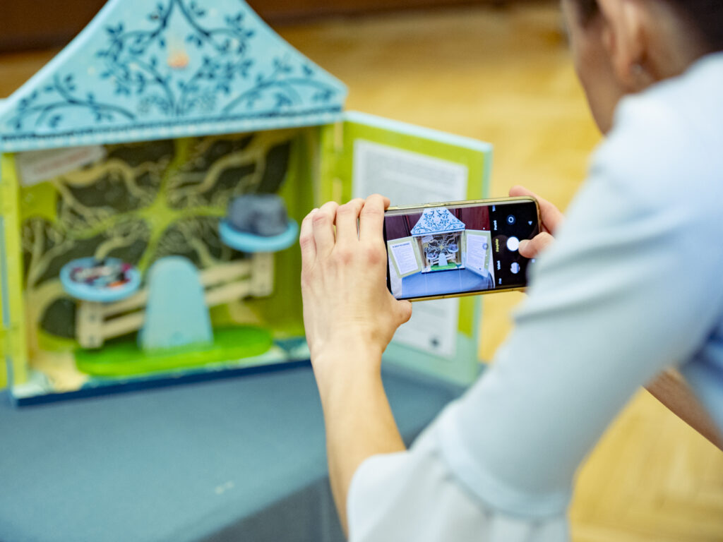 Egy fiatal a telefonjával fotóz egy asztalon álló, színes, miniatűr diorámát. A kis makett ház- vagy színpadszerű szerkezet, kék és zöld mintákkal díszítve. A belső térben apró tárgyak – például egy pad és egy kalap – láthatók. A jelenet a kreatív alkotást és annak dokumentálását mutatja be.
