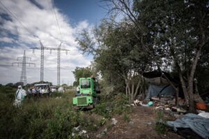 Fák között kialakított, ponyvákból és műanyag fóliákból épített ideiglenes táborhely egy bozótos területen. A földön szétszórt hulladék, háztartási tárgyak és különféle használati eszközök láthatók. A helyszín mellett egy zöld munkagép áll, a háttérben több önkormányzati és hatósági munkatárs figyeli a felszámolási munkálatokat. A terület felett nagyfeszültségű elektromos vezetékek húzódnak. Az önkormányzat július 21-én felszámolta az Erdőkerülő utca közelében kialakított illegális hajléktalantanyákat.