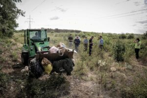 Bozótos, füves területen egy zöld munkagép a felszámolt táborhelyről összegyűjtött hulladékot és használati tárgyakat szállít. A háttérben több önkormányzati és hatósági munkatárs figyelemmel kíséri a munkálatokat. A terület felett nagyfeszültségű vezetékek húzódnak. A kép az Erdőkerülő utca közelében kialakított illegális hajléktalantanyák július 21-i felszámolásának egyik mozzanatát mutatja.