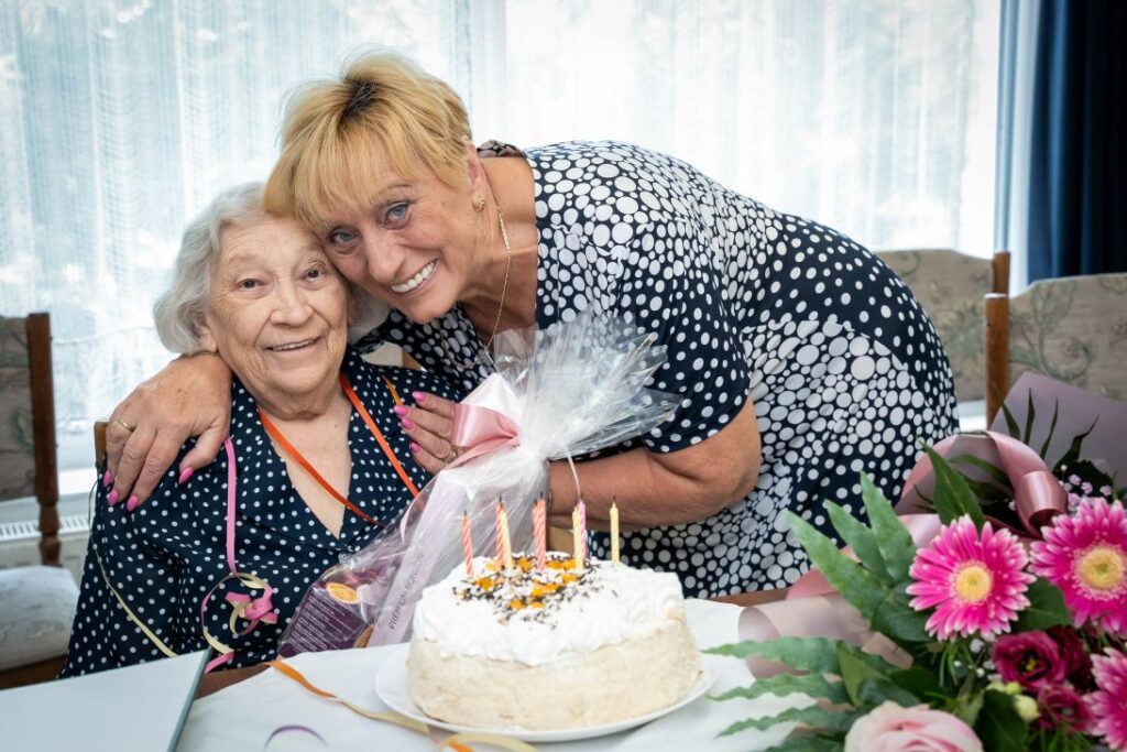 Szepesi Lajosné 90. születésnapjának ünnepségén az Olajág Otthonok Bánkút utcai intézményében. A szépkorú ünnepelt és egy látogató egy asztal mellett ülnek, előttük gyertyákkal díszített születésnapi torta és virágcsokor látható. Az eseményt a XV. kerületi önkormányzat köszöntése alkalmából rendezték.