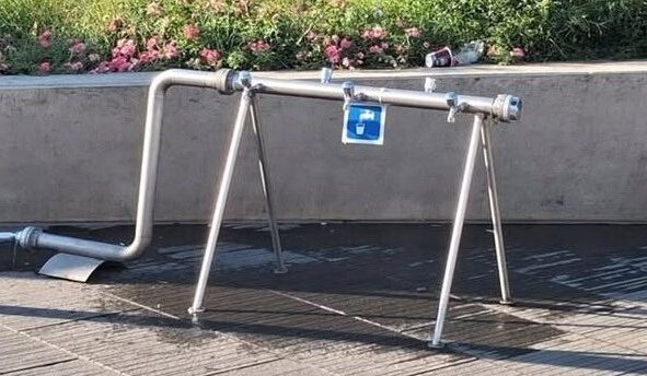 A képen egy ideiglenes, fémből készült ivókút látható A csapállvány négy lábon áll, rajta jól felismerhető az ivóvíz nemzetközi piktogramja. A szerkezet alatt nedves a burkolat, ami arra utal, hogy a kutat nemrég használták.