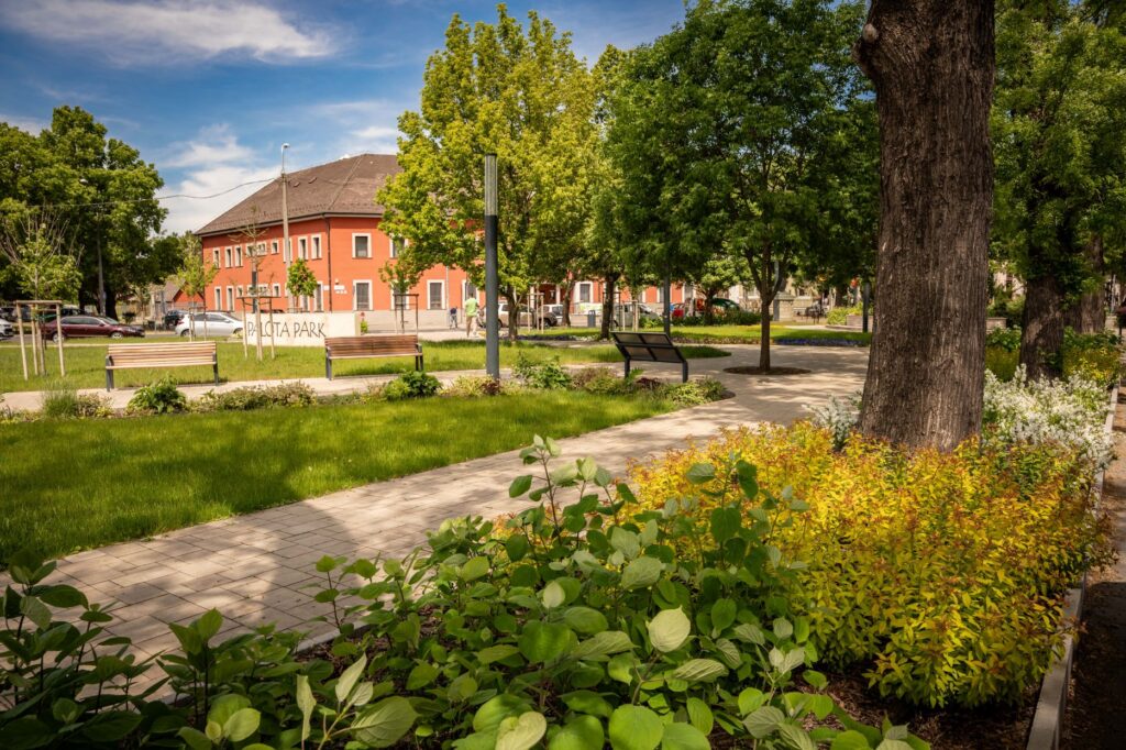A XV. kerületi Rákospalotán található Palota Park részlete. A gondozott közparkban kanyargó térköves sétányok, padok, virágágyások és lombos fák láthatók. Az előtérben díszcserjék és egy idős fa törzse, a háttérben a „Palota Park” felirat és egy narancsszínű, kétszintes épület látható. Napos időben készült felvétel, sok zöld növényzettel és rendezett parkkörnyezettel.