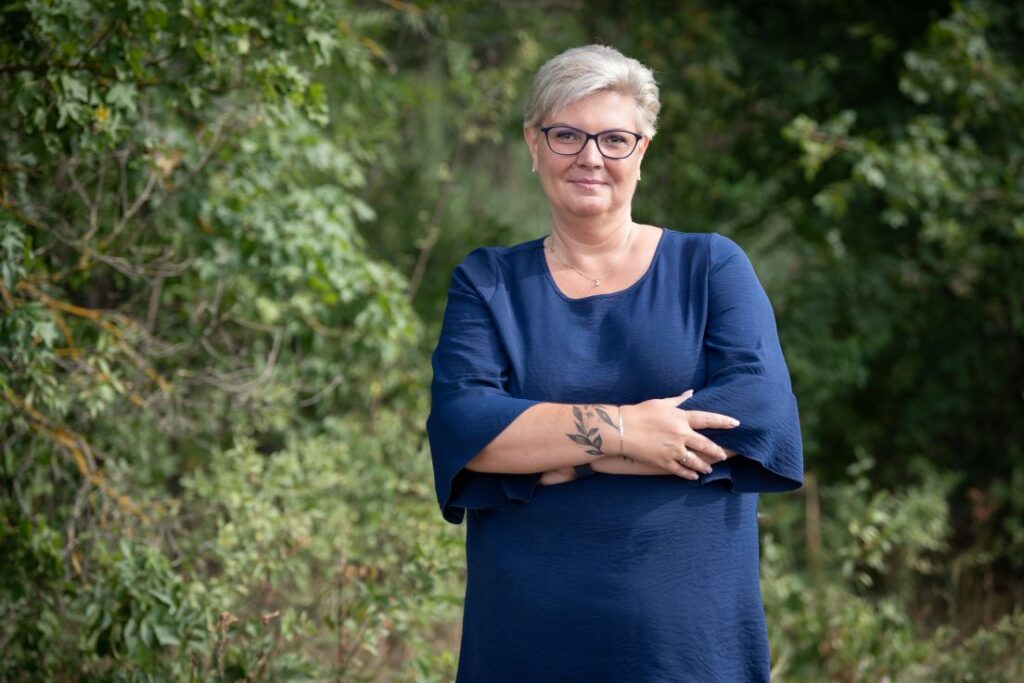Cserdiné Németh Angéla áll egy szabadtéri, zöld növényzettel körülvett helyszínen. Sötétkék, háromnegyedes ujjú ruhát visel és szemüveget hord. Karjait maga előtt összefonja. A háttérben fák és bokrok láthatók, amelyek életlenül jelennek meg a portréfotón. A felvétel természetes fényben készült.