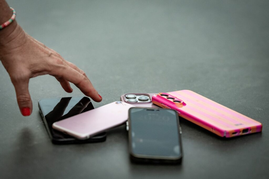 Egy asztalon több használt mobiltelefon fekszik egymásra helyezve. A készülékek között fekete, rózsaszín és pink színű telefonok láthatók, néhányuk tokban van. A kép bal oldalán egy kéz nyúl a telefonok felé. A háttér egyszínű, elmosódott, így a fókusz a régi mobilkészülékeken van. A fotó a használt telefonok gyűjtésének és újrahasznosításának témáját szemlélteti.