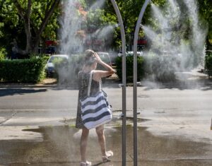 Az Újpalotán működő párakapu egy forró nyári napon finom vízpermetet bocsát ki. A képen egy járókelő halad át a két íves fémszerkezet között, miközben a permet hűsíti a környezetet. A háttérben fák, parkoló autók és egy többszintes lakóépület látható. A napsütésben a vízpára jól kirajzolódik a zöld növényzettel övezett városi környezetben.